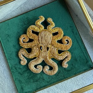 May include: A gold octopus brooch featuring intricate embroidery and beadwork. The octopus has a textured metallic thread body, eight curled tentacles adorned with small gold beads, and two prominent black oval-shaped eyes, centred on a square green velvet base.