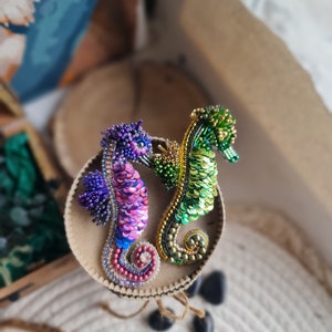 Puede incluir: Dos broches de caballito de mar hechos con cuentas, uno verde y dorado, el otro morado y rosa, sobre una superficie circular marr&oacute;n.