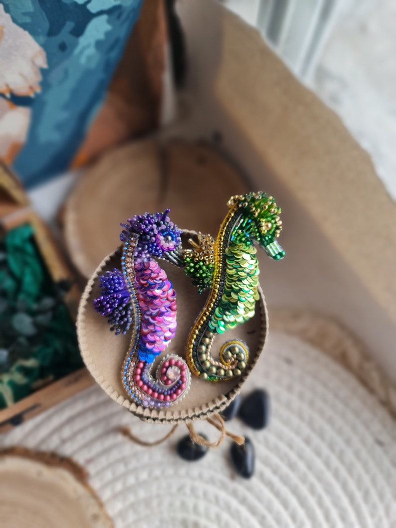 Puede incluir: Dos broches de caballito de mar hechos con cuentas, uno verde y dorado, el otro morado y rosa, sobre una superficie circular marr&oacute;n.