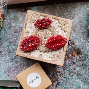 以下が含まれることがあります： 木製の表面に、赤いラインストーンでできた唇の形のブローチが3つあります。ブローチは、彫刻が施された小さな木製の箱に入っています。箱は、グレーのざらざらした表面の上に置かれています。