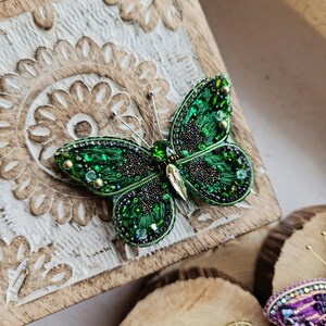 Op de afbeelding: Een groene en zwarte kralen vlinder broche met iriserende vleugels. De broche ligt op een houten oppervlak.