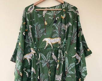 May include: A green, V-neck kaftan dress with a jungle-themed print. The pattern includes zebras, leopards, toucans, and palm trees. The dress is hanging on a wooden hanger.