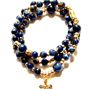 May include: A three-strand beaded bracelet featuring dark blue and gold beads. The bracelet has a gold-colored cross pendant and a gold-colored charm. The beads are a mix of solid and speckled blue.