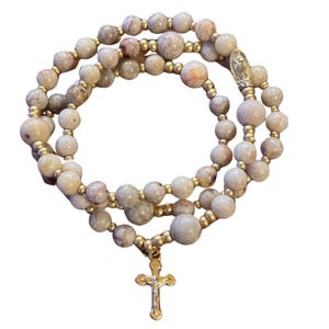 Könnte beinhalten: Ein Rosenkranz mit hellbeige und rosa Steinperlen, verbunden durch goldfarbenes Metall. Der Rosenkranz hat einen goldfarbenen Kreuzanhänger und einen kleinen Anhänger mit einer religiösen Figur.