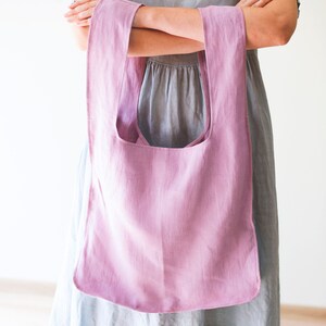 May include: A light pink linen tote bag with a wide, open top and a single strap. The bag is held by a person wearing a light blue dress.