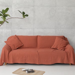 Könnte beinhalten: Ein terracottafarbener Sofaüberwurf aus Leinen mit zwei passenden Kissen. Der Überwurf ist über ein Sofa mit einer hellgrauen Betonwand im Hintergrund gelegt.