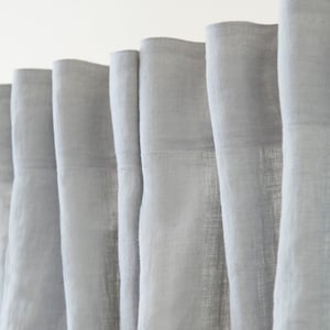 Puede incluir: Primer plano de un conjunto de cortinas de lino gris claro colgadas en una ventana. Las cortinas están hechas de una tela suave y texturizada y tienen un brillo sutil.