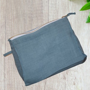 May include: A gray linen zippered pouch with a gold zipper. The pouch is lying on a light wood surface.