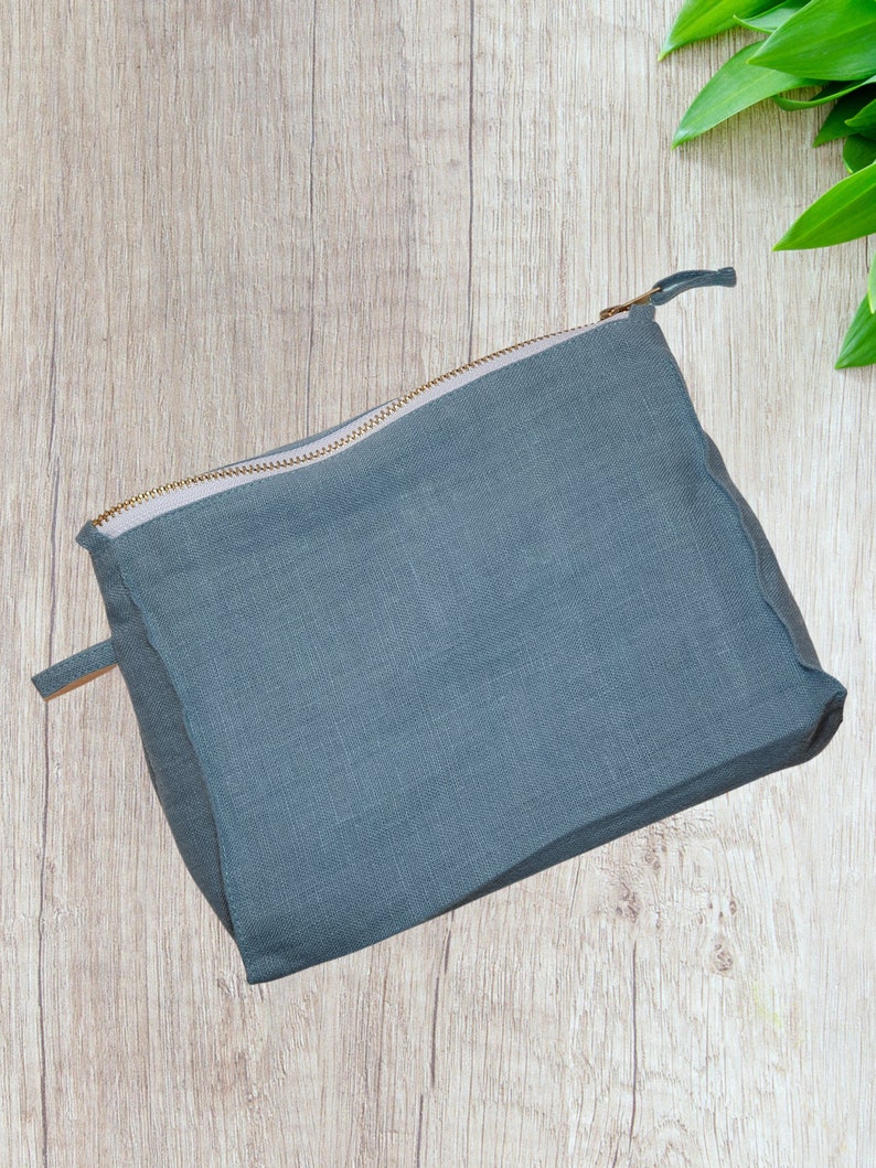 May include: A gray linen zippered pouch with a gold zipper. The pouch is lying on a light wood surface.