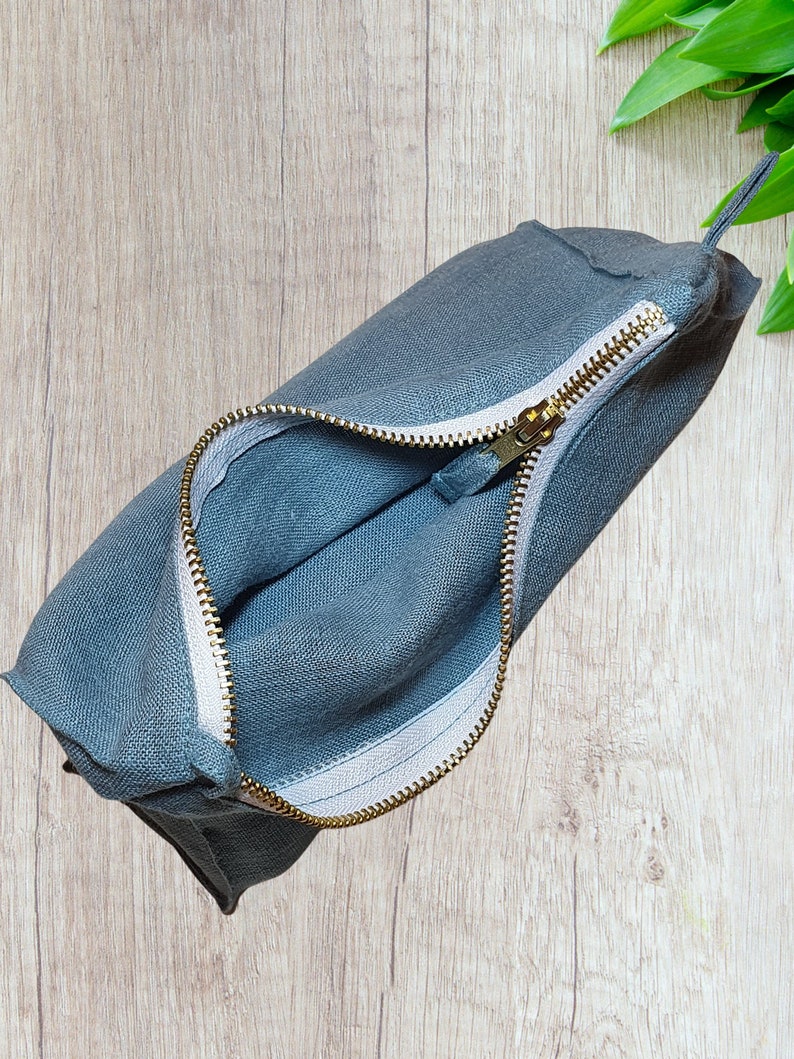 May include: A gray linen zippered pouch with a gold zipper. The pouch is open and the lining is white.