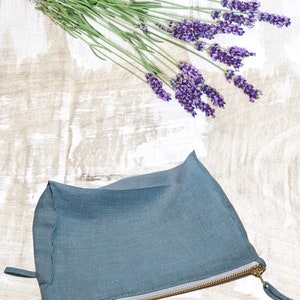 May include: A teal linen zippered pouch on a whitewashed wooden surface with a sprig of lavender flowers.