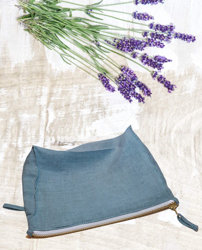 May include: A teal linen zippered pouch on a whitewashed wooden surface with a sprig of lavender flowers.