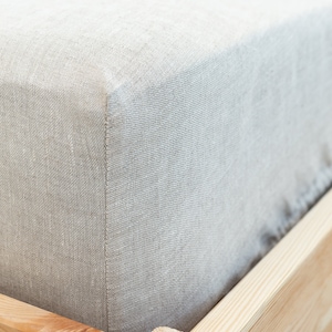 Puede incluir: Primer plano de una sábana bajera de lino gris claro sobre un marco de cama de madera. La sábana es lisa y sin arrugas, con una textura sutil.
