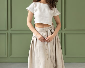 May include: A woman wearing a white linen crop top and a beige linen skirt with a button closure and a tie belt. The skirt is midi length and has a pleated design. The woman is standing in front of a green wall.