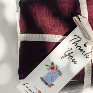May include: A stack of burgundy, gray, and white linen fabric is tied with a white ribbon and a gift tag. The tag has a hand-drawn illustration of a spool of thread with flowers and the text "Thank You" and "with Linen Couture a boutique shop".