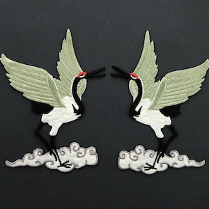 May include: Two embroidered iron-on patches of white cranes with black and red accents, standing on gray clouds. The cranes are facing each other with their wings spread.