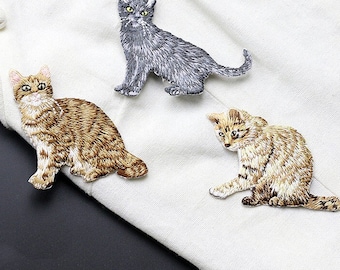 May include: Three embroidered iron-on patches featuring different cat designs. The patches are gray, brown, and tan.