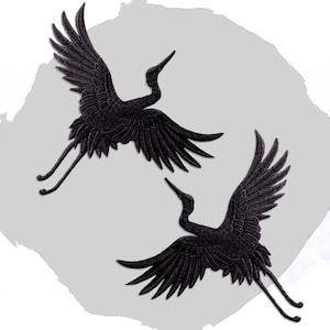 May include: Two black crane appliques in flight, with outstretched wings and detailed feather textures. The birds are positioned against a soft gray and white background, creating a sense of movement and elegance.