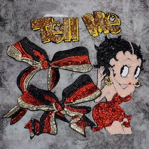 Può includere: Toppe ricamate raffiguranti una donna dei cartoni animati in un abito di paillettes rosse, capelli neri e orecchini d'oro. Due toppe a forma di fiocco di paillettes in nero, rosso e oro. Le parole "Tell Me" sono scritte con paillettes dorate.