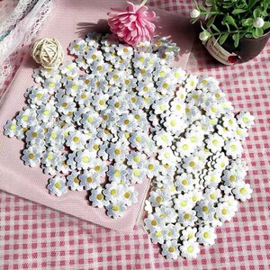 Peut inclure: Un grand nombre de patchs en tissu de forme de marguerite blanche avec des centres jaunes. Les patchs sont disposés de manière aléatoire sur une surface à carreaux rose et blanche.