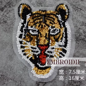 May include: A tiger head patch crafted with gold, black, and white sequins and beads on a mesh backing. The design features a red sequined tongue and measures 7.5 cm in width and 16 cm in height. The text EMBROIDII is visible on the image.