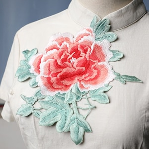 May include: A white fabric with a large embroidered pink and white flower with green leaves.
