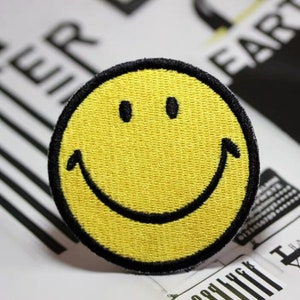 May include: Yellow smiley face embroidered patch with a black outline. The patch is approximately 5 cm in diameter.