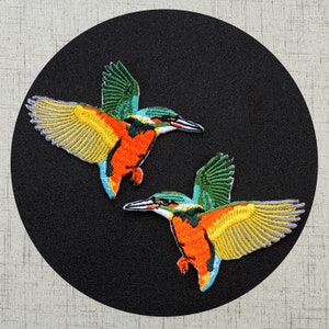 May include: Two embroidered iron-on patches of kingfishers in flight. The birds are green, blue, yellow, orange, and gray.