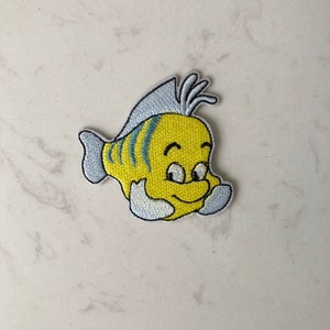 Könnte beinhalten: Ein gestickter Bügel-Aufnäher von Flounder aus dem Disney-Film Arielle, die Meerjungfrau. Flounder ist ein kleiner Fisch mit einem großen Lächeln und schaut nach rechts.