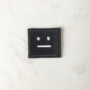 May include: Black embroidered patch with a white face featuring two dots for eyes and a straight line for a mouth.