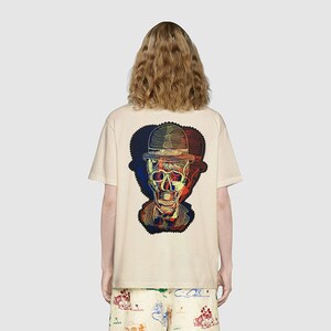 May include: A beige short-sleeved t-shirt featuring a large, multi-colored graphic on the back. The design depicts a skull wearing a bowler hat, rendered in overlapping red, blue, and yellow lines that create a vibrant, layered visual effect.