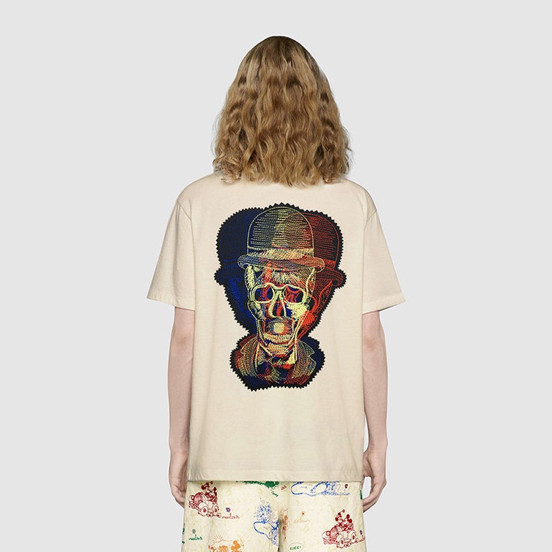 May include: A beige short-sleeved t-shirt featuring a large, multi-colored graphic on the back. The design depicts a skull wearing a bowler hat, rendered in overlapping red, blue, and yellow lines that create a vibrant, layered visual effect.