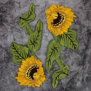 May include: Two embroidered sunflowers with green leaves on a gray background. The sunflowers are yellow with brown centers. The leaves are a dark green.