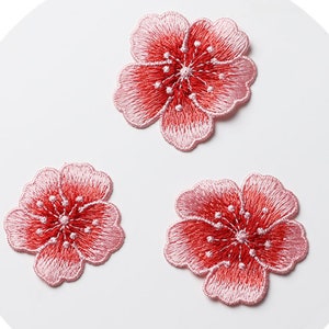 May include: Three pink and red embroidered flower patches. The flowers have five petals and a center with a small circle and several small dots.