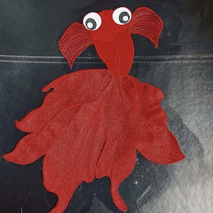 Könnte beinhalten: Ein roter Filzfisch mit großen weißen Augen und einem lächelnden Mund. Der Fisch hat ein detailliertes Design mit überlappenden Schuppen und einem fließenden Schwanz.