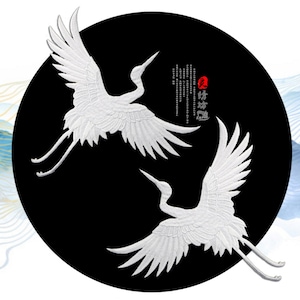 May include: Two white embroidered cranes on a black circular background. The cranes are facing opposite directions and their wings are spread out. The background has Chinese calligraphy in black and red.