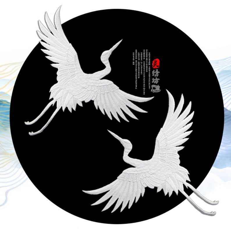 May include: Two white embroidered cranes on a black circular background. The cranes are facing opposite directions and their wings are spread out. The background has Chinese calligraphy in black and red.