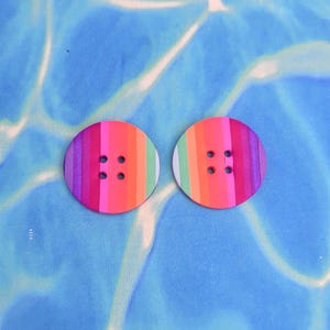 May include: Two circular buttons with a colourful rainbow stripe pattern. Each button has vertical stripes in pink, orange, green, and purple. The buttons have four holes for sewing and are set against a blue background.