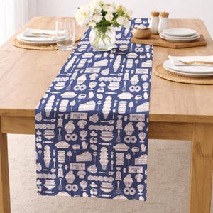 Op de afbeelding: Een marineblauwe tafelloper met een wit patroon van bakgerelateerde illustraties, waaronder taarten, ijs en keukengereedschap. De loper ligt op een houten tafel, met een vaas met witte bloemen en servies.
