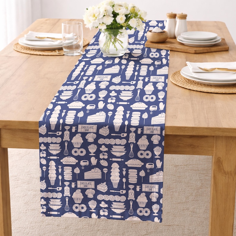 Op de afbeelding: Een marineblauwe tafelloper met een wit patroon van bakgerelateerde illustraties, waaronder taarten, ijs en keukengereedschap. De loper ligt op een houten tafel, met een vaas met witte bloemen en servies.