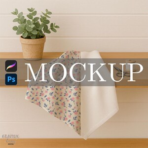 May include: A folded fabric with a floral pattern of pink flowers and blue leaves, alongside a plain white fabric. The word "MOCKUP" is overlaid on the fabric. A small potted plant, scissors, and a thread spool are also visible.