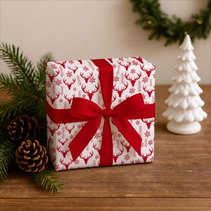 Pu&ograve; includere: Un regalo avvolto in carta bianca con un motivo di cervi e fiocchi di neve rossi, legato con un nastro e un fiocco rossi. Poggia su una superficie di legno, con pigne, un piccolo albero di Natale bianco e vegetazione sullo sfondo.