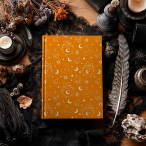 May include: An orange journal with a white Halloween-themed pattern featuring witch hats, cauldrons, moons, and stars. The journal is surrounded by a black lace cloth, a feather, and other decorative items, creating a mystical aesthetic.