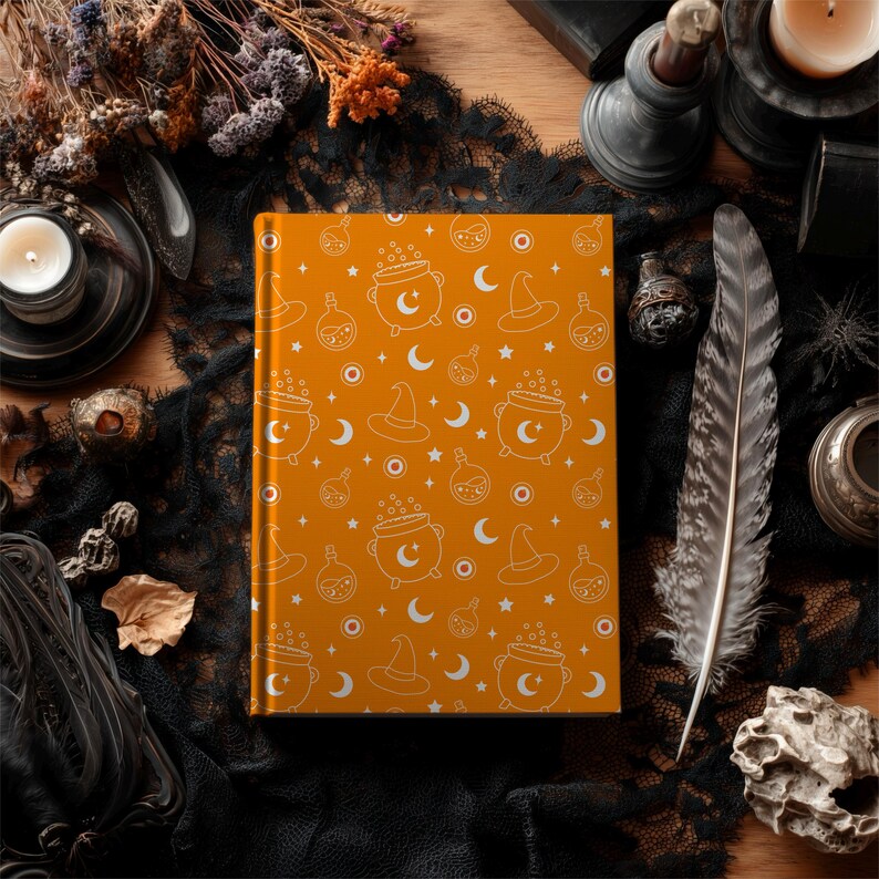 May include: An orange journal with a white Halloween-themed pattern featuring witch hats, cauldrons, moons, and stars. The journal is surrounded by a black lace cloth, a feather, and other decorative items, creating a mystical aesthetic.