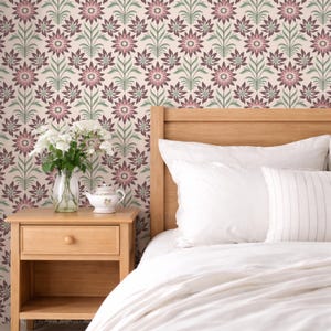 Op de afbeelding: Een slaapkamer met een houten nachtkastje, een bed met wit beddengoed en bloemenbehang. Het behang heeft een herhalend patroon van roze en bordeauxrode bloemen met groene stelen tegen een cr&egrave;mekleurige achtergrond. Een vaas met witte bloemen en een theepot staan op het nachtkastje.