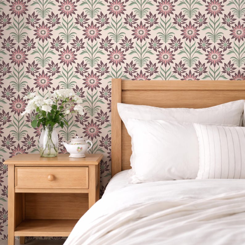 Op de afbeelding: Een slaapkamer met een houten nachtkastje, een bed met wit beddengoed en bloemenbehang. Het behang heeft een herhalend patroon van roze en bordeauxrode bloemen met groene stelen tegen een cr&egrave;mekleurige achtergrond. Een vaas met witte bloemen en een theepot staan op het nachtkastje.