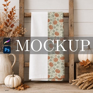 May include: A white kitchen towel featuring a vertical panel with a repeating pattern of orange pumpkins and green leaves on a light background. The towel is draped over a wooden ladder, with the word MOCKUP overlaid in large white text.