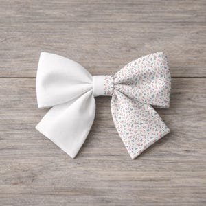 May include: A white fabric bow with a floral patterned accent. The bow features a solid white center and ends, with a section of fabric printed with small pink and blue flowers on a cream background. The bow is set against a wooden surface.