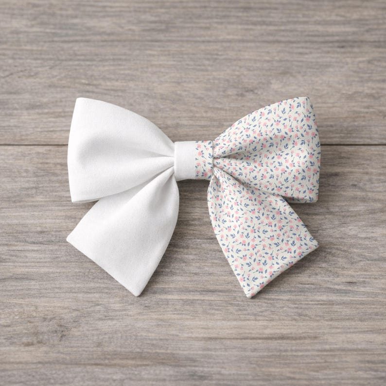 May include: A white fabric bow with a floral patterned accent. The bow features a solid white center and ends, with a section of fabric printed with small pink and blue flowers on a cream background. The bow is set against a wooden surface.