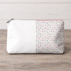 May include: A white zippered pouch with a floral pattern on the right side. The pattern features small pink flowers and blue leaves on a cream background. The pouch has a silver zipper.